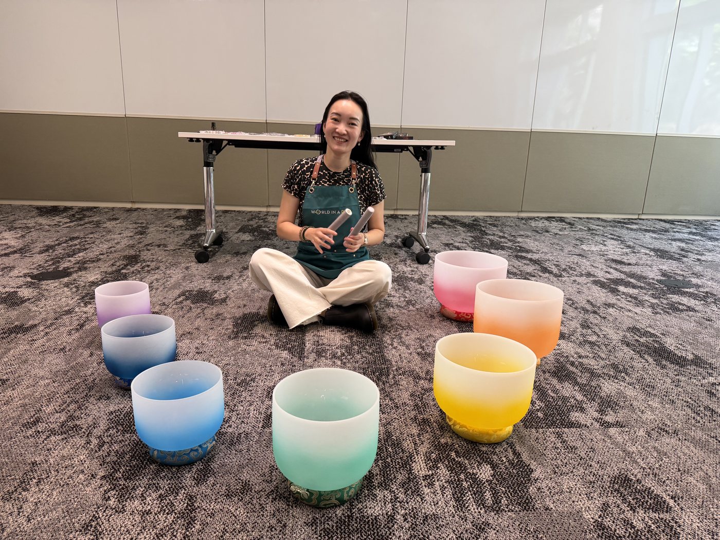 A World in a Dish instructor seated with a circle of colorful crystal singing bowls at an off-site sound bath session